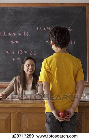 Stock Photo - Schoolboy (12-13) holding apple behind back with teacher in background. Fotosearch