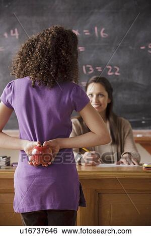 Stock Photograph - Schoolgirl (10-11) holding apple behind back with teacher in background. Fotosearch