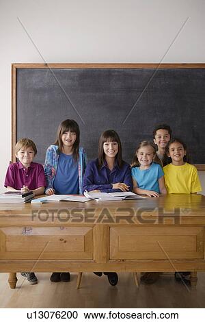Stock Image - Teacher sitting by desk with group of pupils. Fotosearch