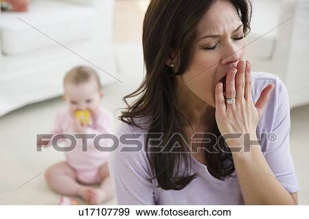 Stock Photo - Tired woman yawning, baby daughter (6-11 months) in background. Fotosearch
