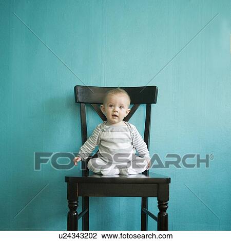 Stock Image - Toddler boy (2-3) sitting on tall chair on blue background. Fotosearch
