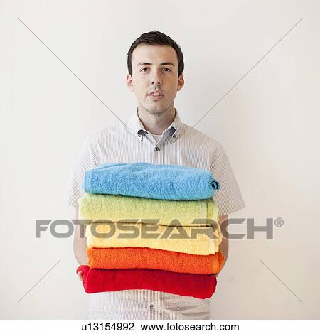 Stock Image - Young man holding stack of multi-coloured towels. Fotosearch