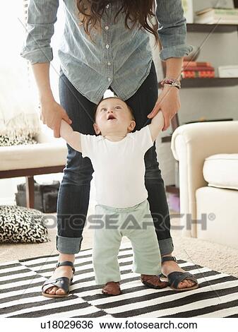 Stock Photograph - Young mother assisting baby boy (6-11 months) in his first steps. Fotosearch