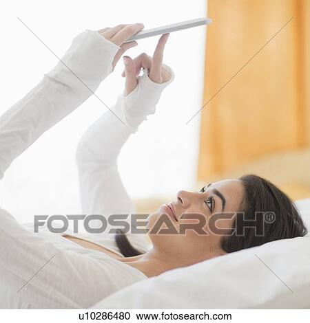 Stock Image - Young woman lying on bed using tablet pc. Fotosearch