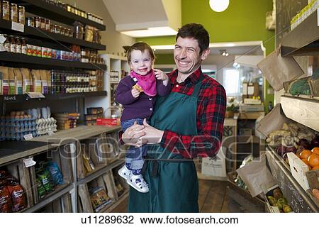 Stock Image - Grocer holding daughter in store. Fotosearch