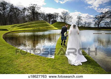 Stock Image - Newlywed couple walking on grassy bridge. Fotosearch
