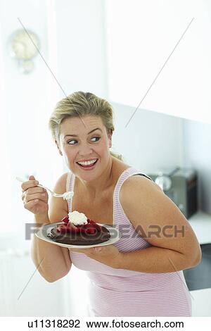 Stock Image - Smiling woman eating cake in kitchen. Fotosearch