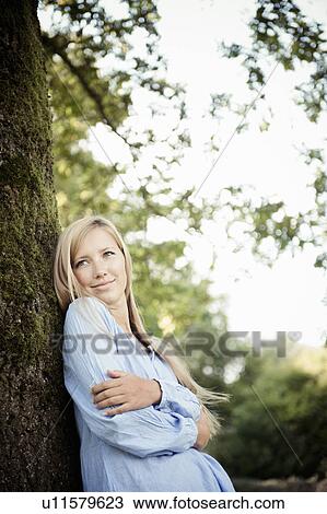 Stock Image - Woman leaning against tree in park. Fotosearch