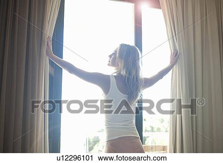 Stock Image - Woman opening curtains in bedroom. Fotosearch