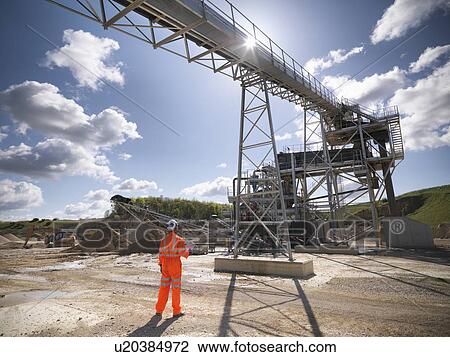 Stock Image - Worker standing under conveyor in quarry. Fotosearch