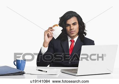 Picture - Businessman eating a burger at the desk. Fotosearch
