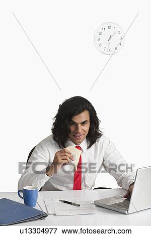 Businessman eating a sandwich while using a laptop at the desk View Large Photo Image Stock Photo - Businessman eating a sandwich while using a laptop at the desk. Fotosearch