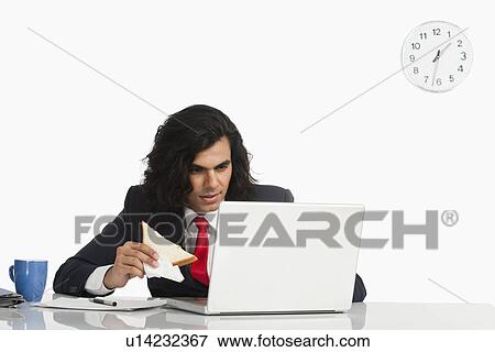 Stock Photo - Businessman eating a sandwich while using a laptop at the desk. Fotosearch