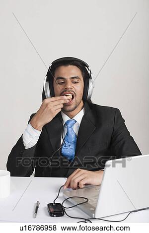 Stock Photo - Businessman eating snacks while listening to headphones at the desk. Fotosearch