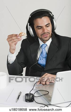 Businessman eating snacks while listening to headphones at the desk View Large Photo Image Stock Photo - Businessman eating snacks while listening to headphones at the desk. Fotosearch