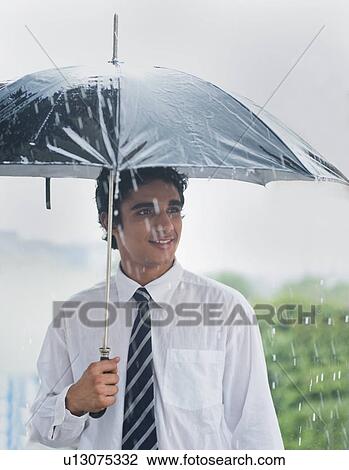Stock Image - Businessman sheltering under umbrella in rain. Fotosearch