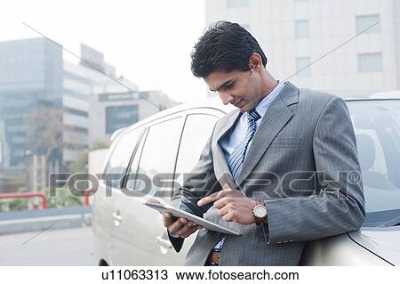 Stock Image - Businessman standing beside a car and using a digital tablet, Gurgaon, Haryana, India. Fotosearch
