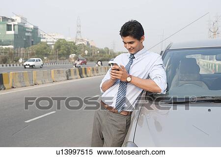 Stock Photography - Businessman standing beside a car and text messaging on a mobile phone, Gurgaon, Haryana, India. Fotosearch