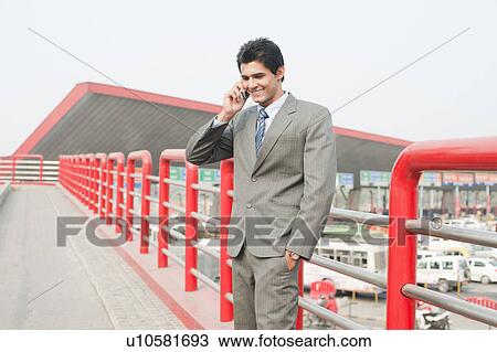 Stock Image - Businessman standing on a footbridge and talking on a mobile phone, Gurgaon, Haryana, India. Fotosearch