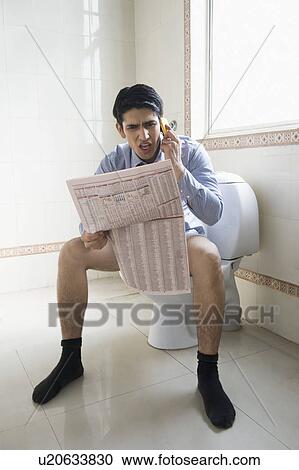 Stock Image - Businessman talking on a mobile phone and reading a newspaper on a toilet seat. Fotosearch