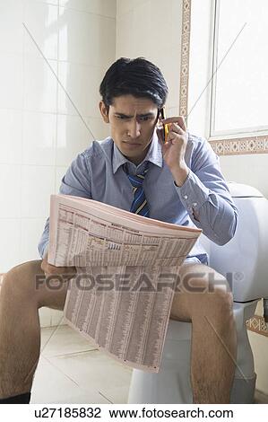 Stock Image - Businessman talking on a mobile phone and reading a newspaper on a toilet seat. Fotosearch