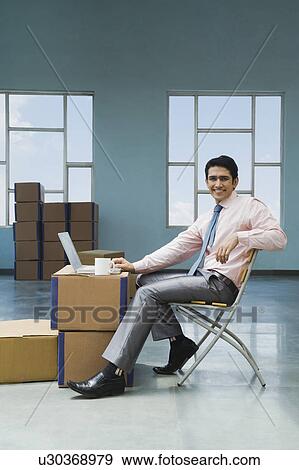 Stock Photo - Businessman using a laptop in a warehouse with cardboard boxes. Fotosearch