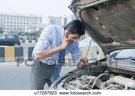 Stock Image - Businessman using a mobile phone near a broken down car, Gurgaon, Haryana, India. Fotosearch