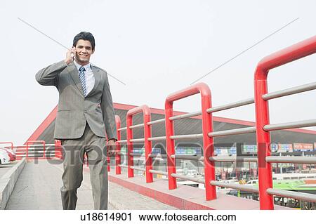 Stock Image - Businessman walking on a footbridge and talking on a mobile phone, Gurgaon, Haryana, India. Fotosearch