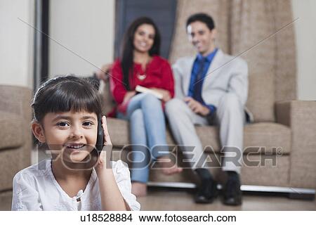 Girl talking on a mobile phone with her parents sitting in the background View Large Photo Image Picture - Girl talking on a mobile phone with her parents sitting in the background. Fotosearch