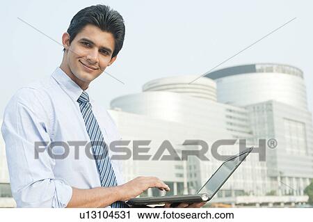 Stock Image - Happy businessman working on a laptop outside an office building, Gurgaon, Haryana, India. Fotosearch