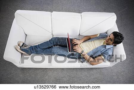 High angle view of a man lying on a couch working on a laptop View Large Photo Image Stock Photograph - High angle view of a man lying on a couch working on a laptop. Fotosearch
