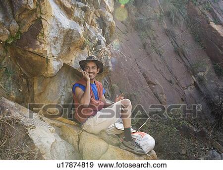 Stock Photo - Hiker sitting on a rock and talking on a mobile phone. Fotosearch
