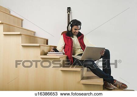 Stock Image - Man listening to music with headphones while using a laptop on a staircase. Fotosearch