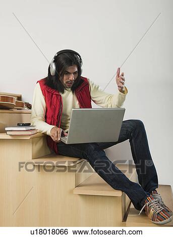 Man listening to music with headphones while using a laptop on a staircase View Large Photo Image Stock Photograph - Man listening to music with headphones while using a laptop on a staircase. Fotosearch