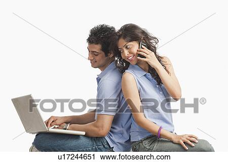 Stock Image - Man using a laptop with his girlfriend talking on a mobile phone. Fotosearch