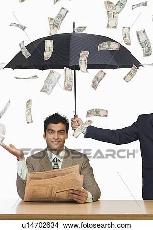 Picture - Money falling on businessman sitting under an umbrella. Fotosearch