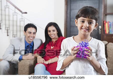Stock Photograph - Portrait of a girl holding a gift with her parents in the background. Fotosearch