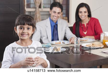 Stock Image - Portrait of a girl holding a glass of milk with her parents in the background. Fotosearch
