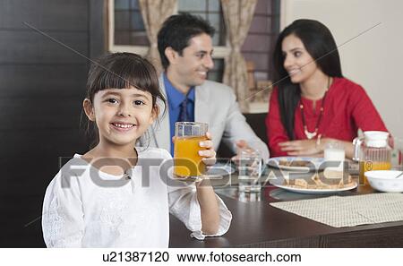 Stock Image - Portrait of a girl holding a glass of milk with her parents in the background. Fotosearch