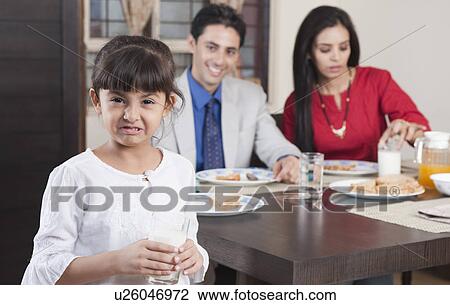 Stock Image - Portrait of a girl holding a glass of milk with her parents in the background. Fotosearch