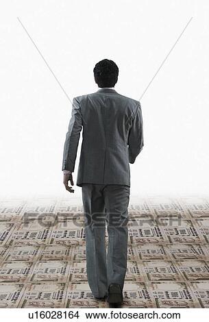 Picture - Rear view of a businessman walking on a path made of Indian paper currency. Fotosearch