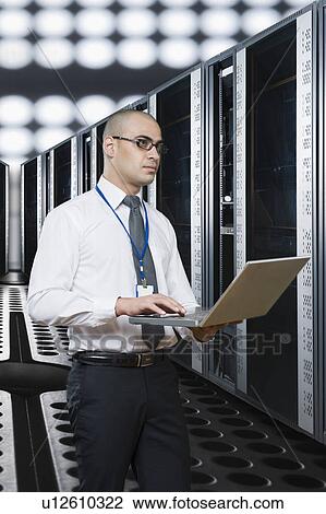 Stock Image - Technician working on a laptop in a server room. Fotosearch