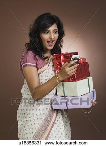 Stock Image - Woman carrying a stack of gift boxes and looking shocked while reading a text message. Fotosearch