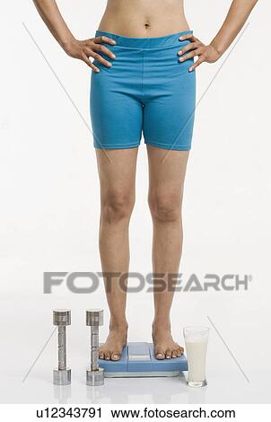 Woman standing on a weight scale with arms akimbo View Large Photo Image Stock Image - Woman standing on a weight scale with arms akimbo. Fotosearch