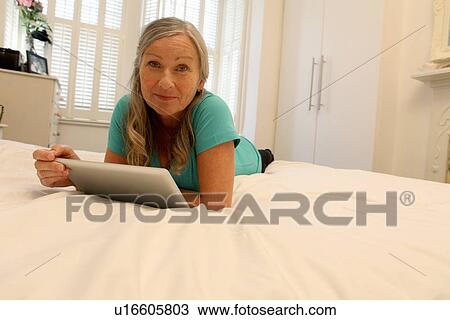 Stock Image - Older woman using tablet computer on bed. Fotosearch