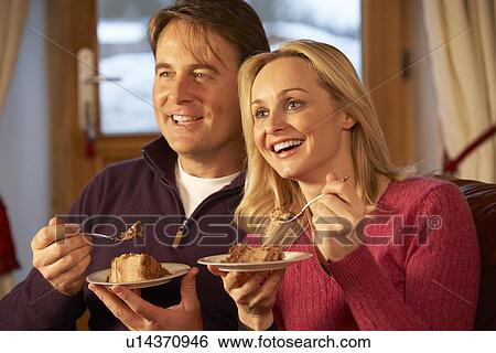 Stock Photograph - Couple Enjoying Slice Of Cake Sitting On Sofa Watching TV. Fotosearch