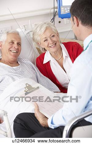 Picture - Doctor talking to senior couple on UK Hospital ward. Fotosearch