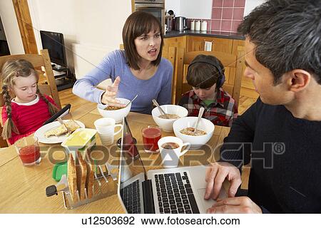 Stock Image - Family Using Gadgets Whilst Eating Breakfast Together In Kitchen. Fotosearch