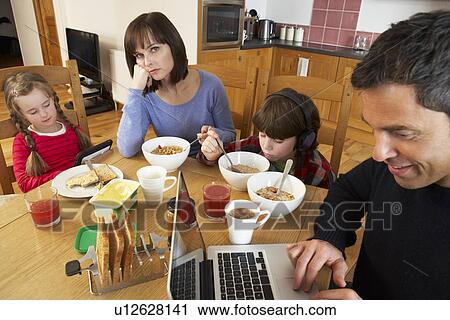 Stock Image - Family Using Gadgets Whilst Eating Breakfast Together In Kitchen. Fotosearch