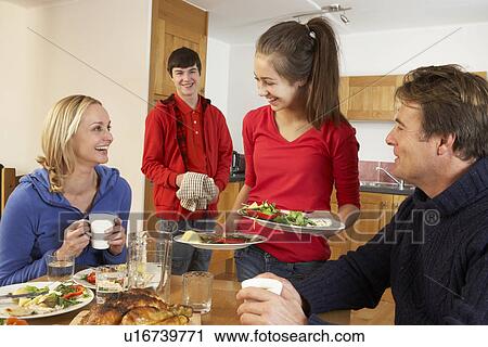 Stock Image - Helpful Teenage Children Serving Food To Parents In Kitchen. Fotosearch